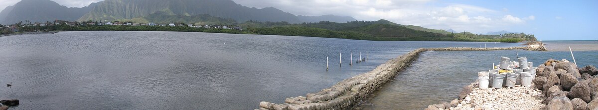 A long stone wall curving across calm shallow water, with green mountains on the far shore.
