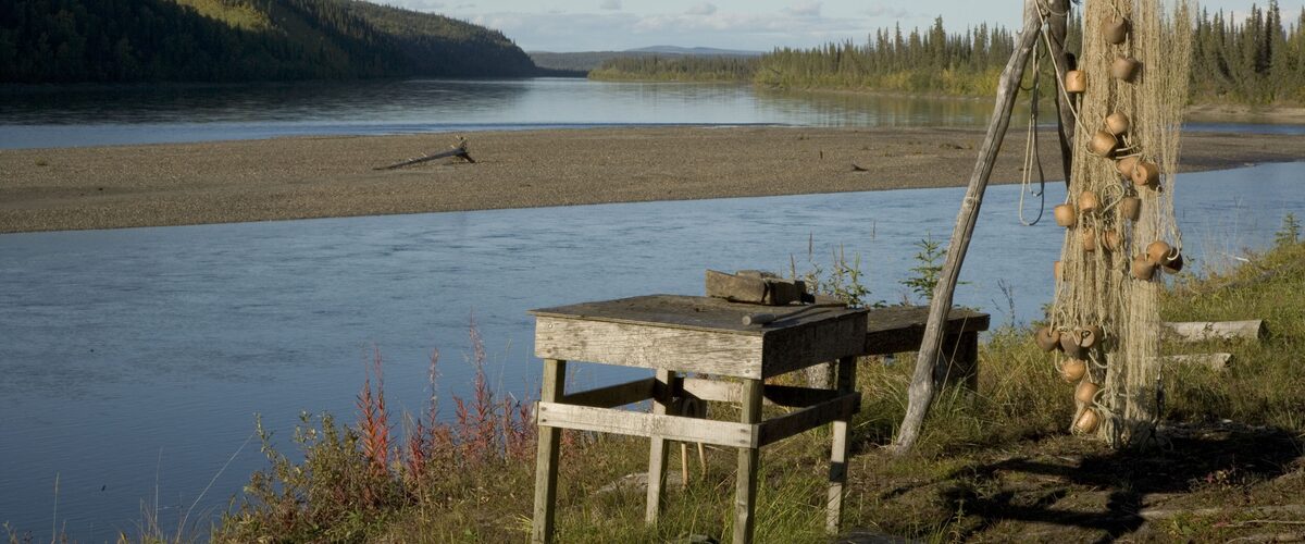 A wooden work table and a fishing net hung on a pole frame on a grassy riverbank, with a wide river, a gravel bar and forested hills beyond.